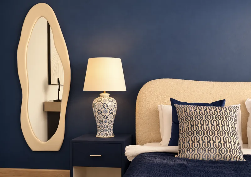 Navy bedroom detail with blue-and-white porcelain lamp, boucle headboard, geometric cushion and cream wavy mirror