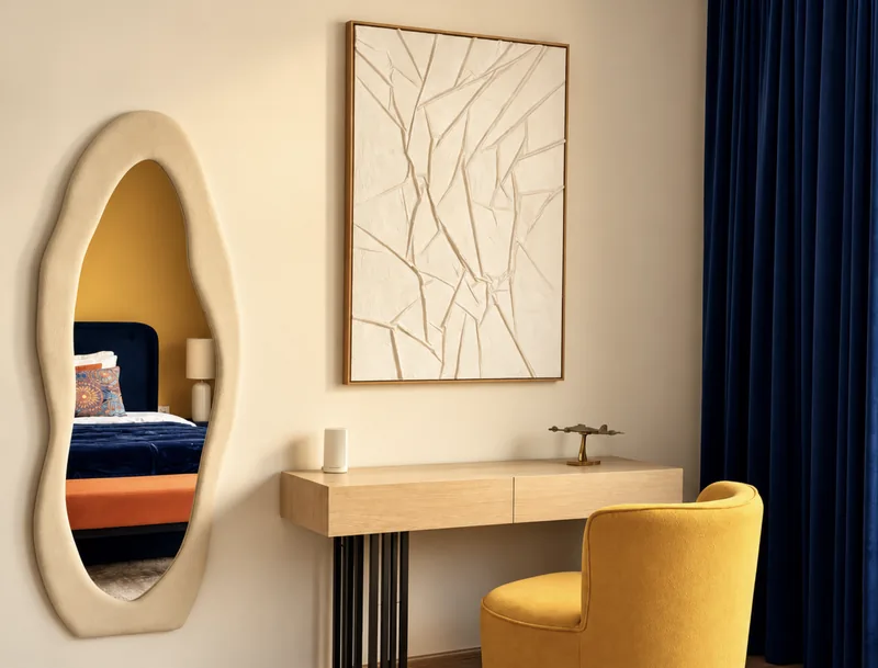 Dressing table with yellow tub chair, textured white artwork and wavy mirror reflecting the yellow bedroom
