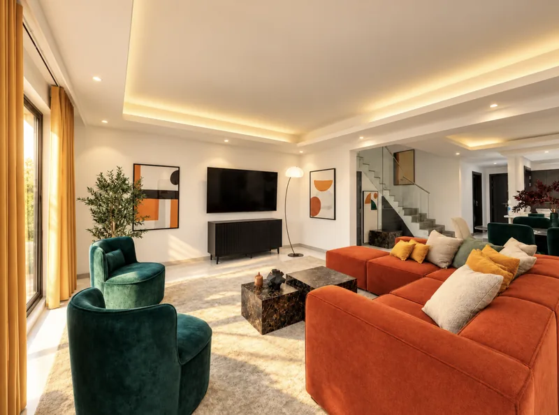 Dunia living room with orange sectional, green velvet armchairs, wall-mounted TV, cove lighting and glass-railed staircase beyond